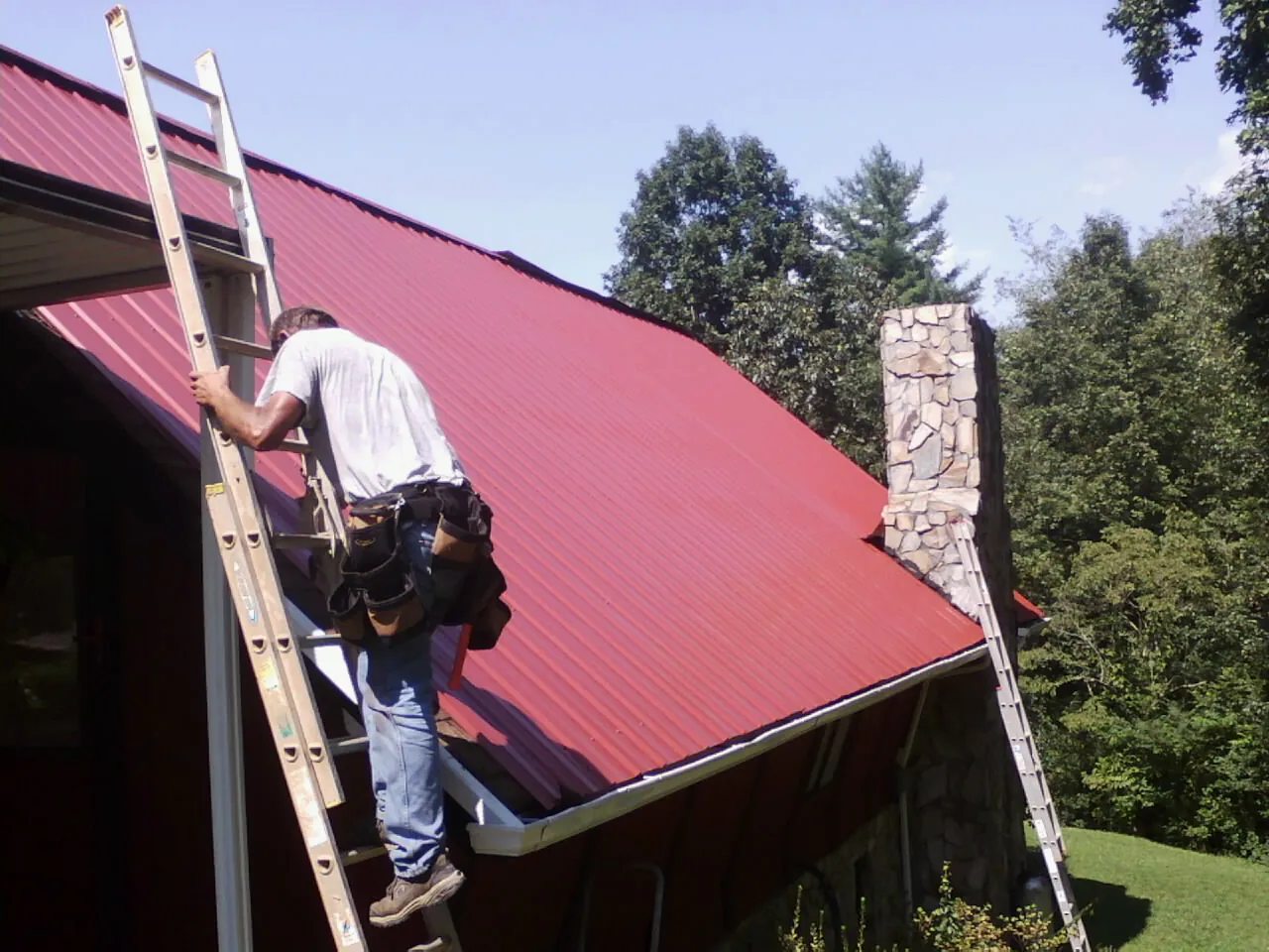 Roofer on a ladder beside a red metal roof and stone chimney — Soffit & Fascia Repair by Roofing Brothers in Pebble Beach